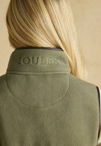 Blondīgs cilvēks, kurš valkā olīvu zaļa flīsa vestu ar izšūtu uzrakstu "JOULES" uz muguras apkakles, fonā ir bēšs fons.