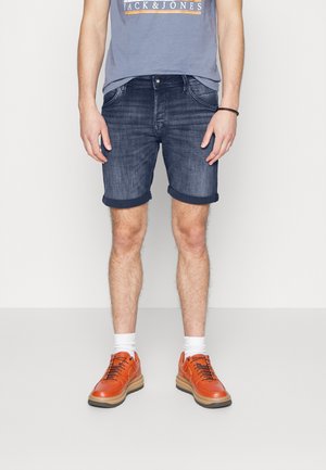 Shorts en denim bleu foncé avec des ourlets roulés, associés à des baskets oranges et des chaussettes blanches. Le haut présente un imprimé logo.