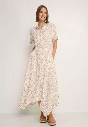 Kurzärmeliges, knöpfbares Kleid in hellbeige mit braunen Polka-Dots, elastischem Bund und asymmetrischem Saum. Das Modell trägt braune Sandalen.