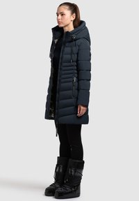 Temno modra puhovka s kapuco, z zadrgo spredaj, stranskimi žepi in prešitim vzorcem. Kombinirana s črnimi legicami in sijočimi črnimi visokimi škornji.