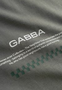 Σκούρο γκρι ύφασμα με το κείμενο "GABBA", μια σύντομη περιγραφική παράγραφο και ένα πράσινο καρό μοτίβο κοντά στο κάτω μέρος.