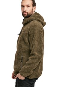 Hoodie en polaire vert olive, avec une surface texturée et douce. Il dispose d'une poche zippée à l'avant et d'un cordon de serrage réglable, offrant une coupe décontractée.
