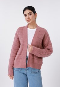 Ροζ πλεκτή ζακέτα με ριμπ υφή και ανοιχτό μπροστινό μέρος. Συνδυασμένη με λευκό τοπ και μπλε τζιν, διαθέτει μακριά μανίκια και άνετη γραμμή.