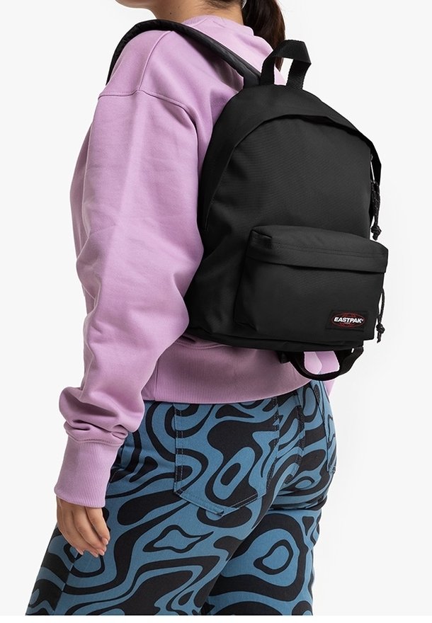 Černý látkový batoh s přední kapsou, nastavitelnými popruhy a logem, nošený přes fialovou mikinu a vzorované modré kalhoty.