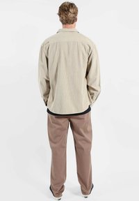 Chemise en velours côtelé beige à rayures verticales, manches longues et ourlet incurvé, associée à un pantalon marron ample. Sous-pull noir visible.