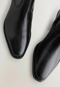 Botas de cuero negro hasta el tobillo con un diseño elegante y de punta afilada. Textura suave con sutiles detalles de costura cerca de los paneles laterales elásticos.