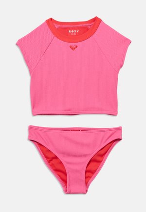 Maillot de bain deux pièces rose texturé avec un haut à manches courtes, doté d'un col rouge et d'un logo en forme de cœur, ainsi que d'un bas de bikini assorti avec une doublure rouge.