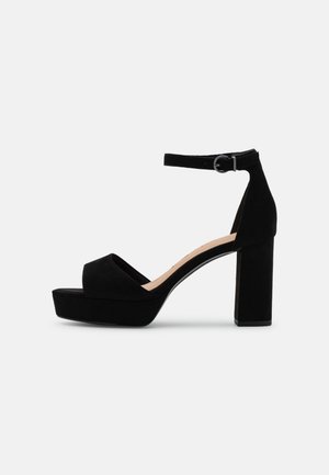 Sandale plateforme en daim noir avec bride à la cheville, talon bloc et bout ouvert, vue de côté sur fond blanc.