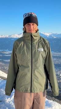 Άτομο που φοράει πράσινο μπουφάν North Face και μαύρο σκουφάκι στέκεται σε χιονισμένο βουνό με χιονισμένες κορυφές και καθαρό γαλάζιο ουρανό στο φόντο.