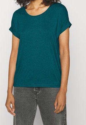 Kvinde iført en teal kortærmet skjorte med opkøbte ærmer og grå jeans, der står mod en ensartet lys baggrund.