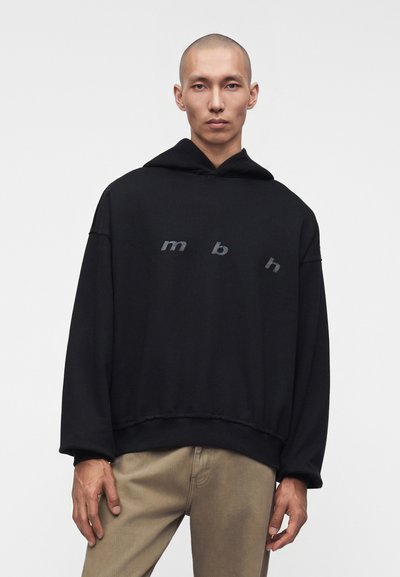 Homme portant un sweat à capuche noir oversize avec des lettres "mbh" sur la poitrine et un pantalon beige, debout devant un fond uni.