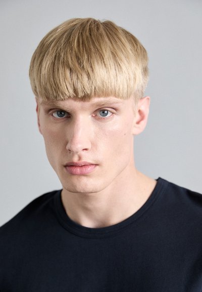 Lys blond hår med rett klipp, blå øyne og naturlig hudtone; iført en ensfarget svart t-skjorte med rund hals mot en nøytral bakgrunn.