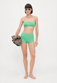 Bikini verde a costine con spalline regolabili, abbinato a pantaloni corti a vita alta. Il modello tiene un asciugamano a righe nere e beige.
