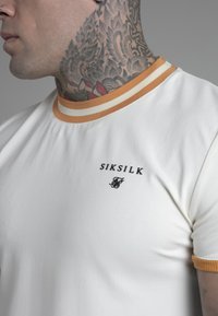 T-shirt bianco a maniche corte con colletto a righe arancione e crema, con il logo nero "Siksilk" sul petto. Texture di cotone liscia.