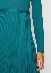 Personne portant une robe bleu sarcelle à manches longues avec jupe plissée et ceinture à la taille, montrée du torse à la main sur un fond uni.