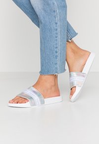 Sandali bianchi con fasce iridescenti che presentano il marchio "TOMMY", abbinati a jeans in denim leggero e scolorito blu.