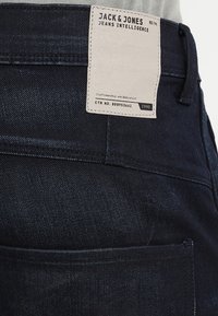 Mörka denimjeans med en strukturerad yta. Har en vit etikett på baksidan med svart text, inklusive varumärkesnamn och skötselråd.