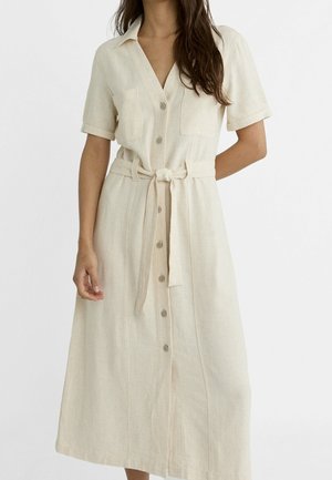 Mujer con vestido beige de manga corta con botones, bolsillos en el pecho y cinturón de tela anudado en la cintura.