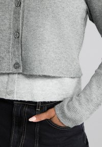 Šedý pletený cardigan s ribbovaným lemem, obsahující kulaté knoflíky a dlouhé rukávy. Nositý přes tmavé džíny s viditelným šitím.