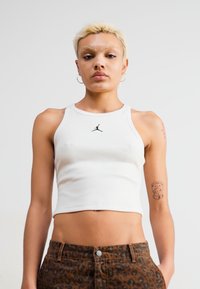 Fehér ujjatlan crop top fekete Jordan logóval, barna leopárdmintás farmerral párosítva. Az anyag simának és testhezállónak tűnik.