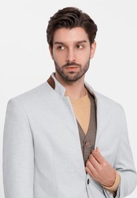 Blazer grigio chiaro con un design strutturato, caratterizzato da un accentuato colletto marrone. Indossato sopra un maglione beige e un gilet a quadri, che mette in mostra trame sovrapposte.