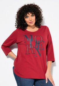 Rode blouse met lange mouwen en een ronde halslijn, voorzien van een decoratieve grafische print van het woord "MODERN" in contrasterende kleuren en texturen.