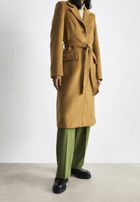 Lång, senapsgul kappa i mjukt tyg med skärp i midjan, två framfickor och jackslag med hack, kombinerad med gröna byxor och svarta skor.