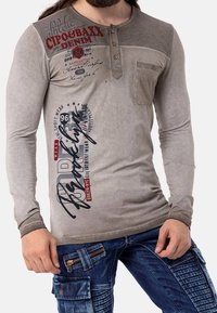Camiseta henley de manga larga gris con botones, con gráficos de texto. Combinada con pantalones cargo de mezclilla azul con cremalleras y detalles texturizados.