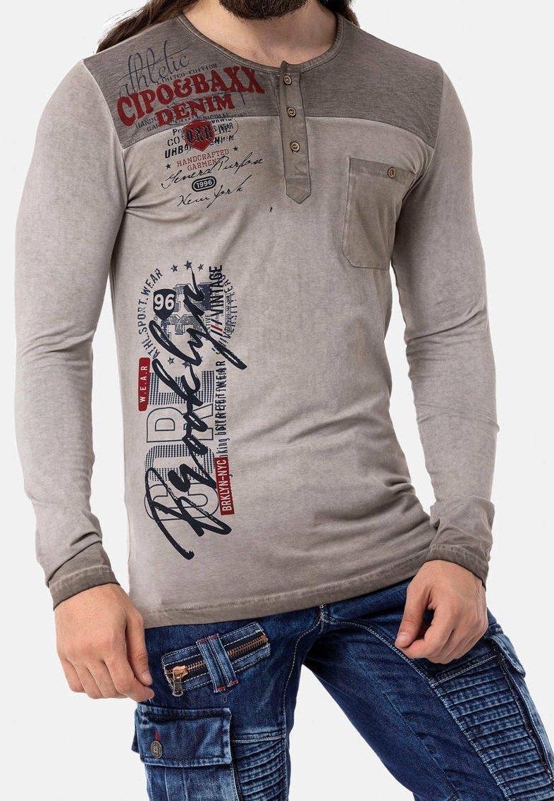 Camiseta henley de manga larga gris con botones, con gráficos de texto. Combinada con pantalones cargo de mezclilla azul con cremalleras y detalles texturizados.