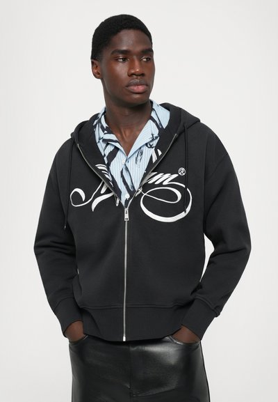 Giovane con le mani in tasca che indossa un hoodie nero con zip sopra una camicia blu a fantasia, su uno sfondo bianco semplice.