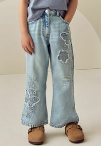 Ljusblå denimjeans med vid passform, försedda med broderade blommor och fjärilsdesigns, samt slitningar vid knäet.