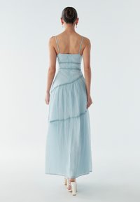 Světle modré maxi šaty s tenkými ramínky, texturovanou látkou a vrstevnatým designem. Mají přiléhavý živůtek a splývavou sukni. Bílé podpatky jsou vidět.