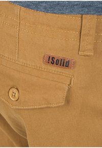 Pantaloni di cotone beige con una tasca posteriore, caratterizzati da un'etichetta in pelle marrone con la scritta "!Solid" stampata in nero. Cuciture e dettagli del bottone visibili.
