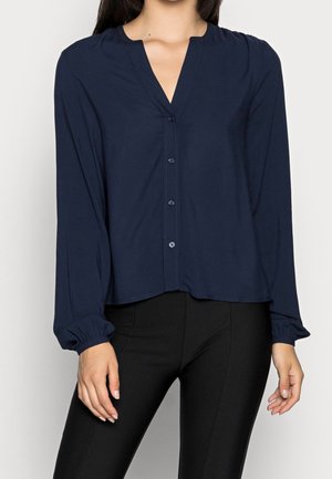 Granatowa bluzka z długim rękawem, dekoltem w serek, zapięciem na guziki oraz marszczonymi mankietami. Materiał jest gładki, o luźnym kroju.