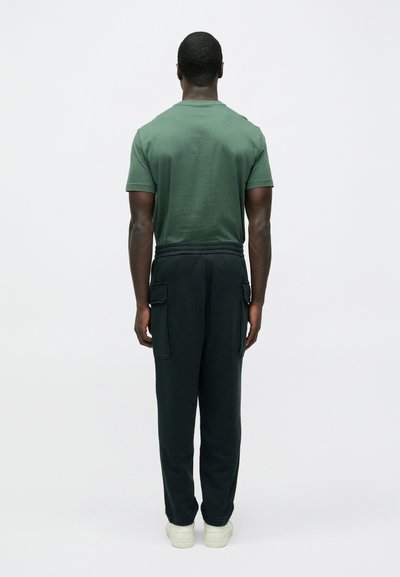 Tmavozelené tričko a čierne tepláky s bočnými vreckami, zobrazené zozadu. Mäkká textúra, voľný strih a elastický pás. Biele tenisky.