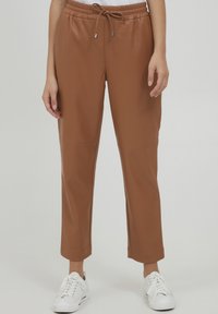 Brune faux læder joggers med elastisk talje, justerbar snor og cropped længde. Båret sammen med hvide sneakers. Glat tekstur.