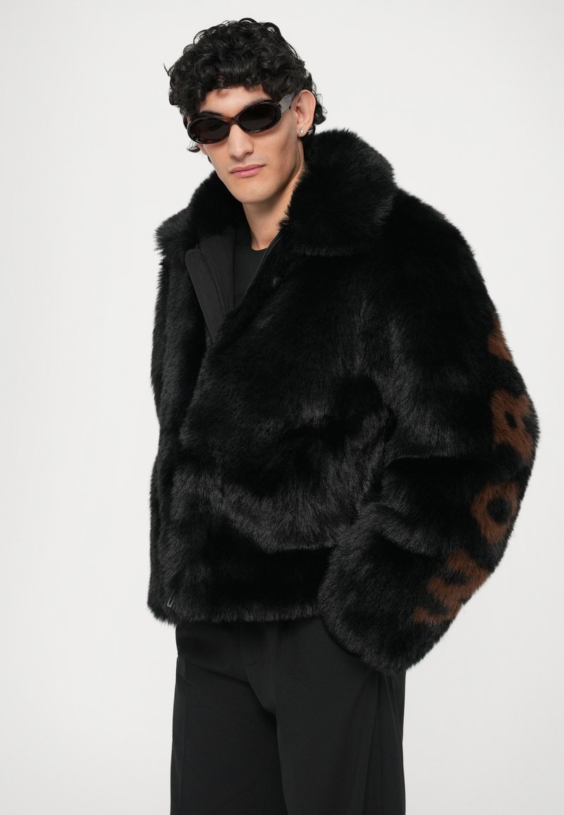 Zwarte nepbontjasje met een brede kraag en een bruine luipaardprintaccent op de linkermouw, gecombineerd met donkere zonnebrillen.