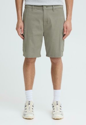 Mand iført olivengrønne cargoshorts, hvide crew-sokker og beige sportssko, stående foran en ensfarvet baggrund.