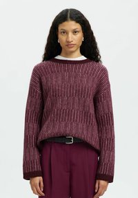 Femme avec de longs cheveux bouclés portant un pull en maille bordeaux texturé et un pantalon bordeaux assorti avec une ceinture noire, faisant face à l'objectif.