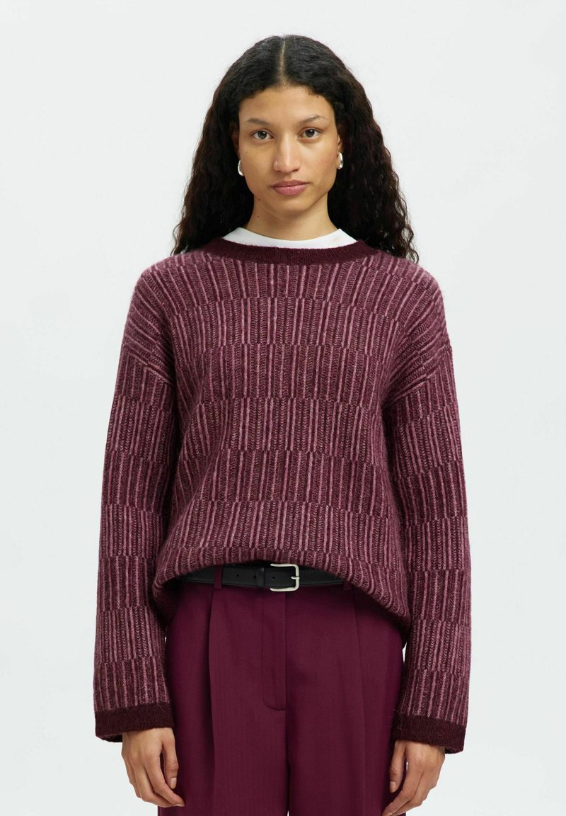 Femme avec de longs cheveux bouclés portant un pull en maille bordeaux texturé et un pantalon bordeaux assorti avec une ceinture noire, faisant face à l'objectif.