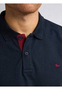 Marineblauwe polo met een bordeauxkleurige knopenlijst, voorzien van twee donkere knopen en een klein rood logo op de borst. Textuurstof.