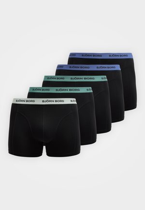 Homme torse nu portant un boxer noir Björn Borg, debout pieds nus sur un tapis clair à côté d'un canapé blanc incurvé contre un mur clair uni.
