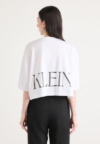 White cropped t-shirt with a large gray print reading "KLEIN" on the back. Oversized fit, short sleeves, soft cotton material.