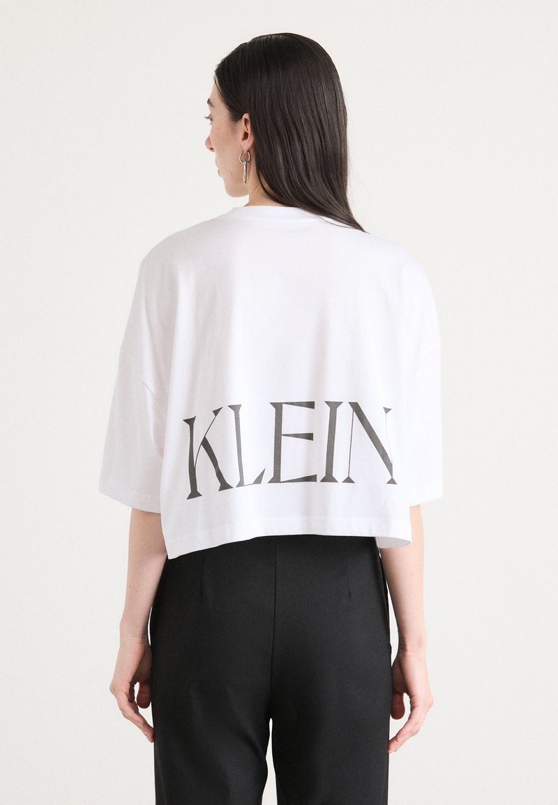 White cropped t-shirt with a large gray print reading "KLEIN" on the back. Oversized fit, short sleeves, soft cotton material.