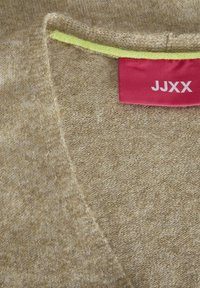 Brun stickad tröja med en mjuk textur, med en v-ringning och en ljusgul accent vid kragen. Röd etikett med texten "JJXX".