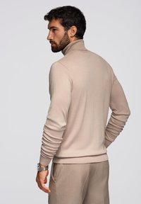 Maglione beige a collo alto in morbido tessuto, design aderente, polsini e orlo a costine. Indossato con pantaloni coordinati, modello di spalle.