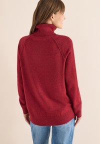 Maglione rosso a collo alto con colletto e polsini a coste, caratterizzato da una texture morbida e una vestibilità ampia, abbinato a jeans azzurri chiari.