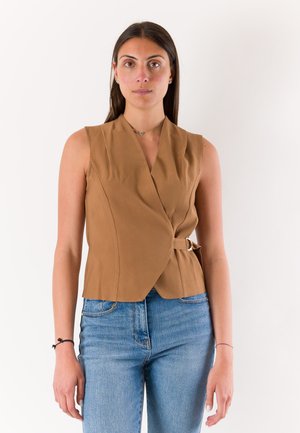 Donna con lunghi capelli scuri che indossa un top avvolgente beige senza maniche e jeans blu, in piedi davanti a uno sfondo bianco uniforme.