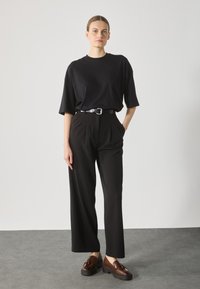 Schwarzes, übergroßes T-Shirt mit lässigem Schnitt. Hoch taillierte schwarze, gefaltete Hose mit geradem Bein. Braune Plateauslöffel mit Quasten-Detail.