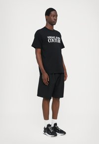 Juodas medvilninis marškinėlis su baltu "VERSACE JEANS COUTURE" logotipu, derinamas su juodais šortais ir juodais sportiniais bateliais su baltais akcentais bei kojinėmis.